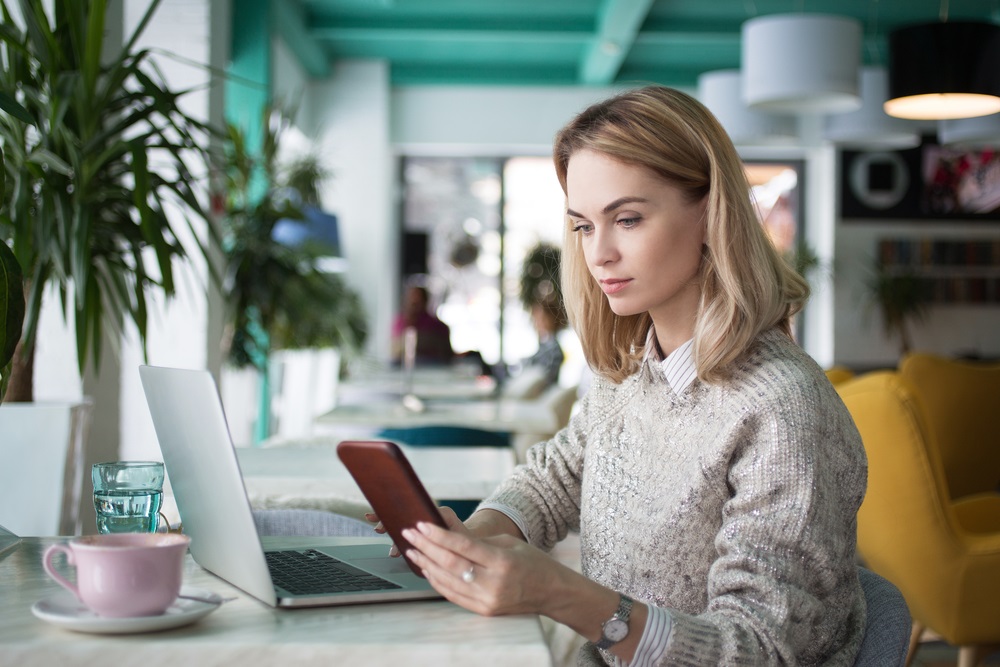 blonde lady checking mobile device in office in front of laptop