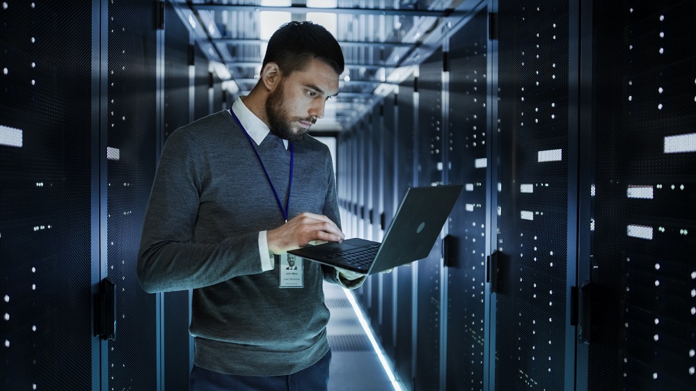 IT support technician checking laptop amongst servers in data center